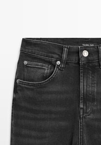 Jeans en denim noir avec une finition texturée, dotés d'une poche avant, d'une fermeture à bouton argenté et d'une étiquette de marque visible à la taille.