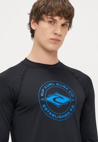 Czarna koszulka z długim rękawem typu rash guard z niebieskim okrągłym logo z napisem "Rip Curl Surf Co" i "Established 69" na klatce piersiowej. Gładka tekstura.