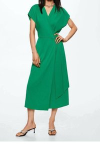 Mujer vistiendo un vestido verde de corte envolvente con mangas cortas y largo medio, combinado con sandalias de tacón negro con tiras de strass.