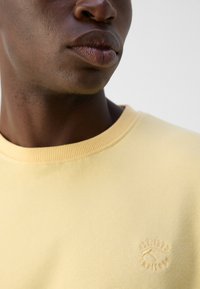 Helle gelbe Sweatshirt mit einem gerippten Halsausschnitt und gesticktem Logo auf der Brust. Das Material wirkt weich und glatt, mit einem lässigen Schnitt.