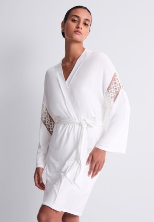 Femme portant une robe portefeuille blanche avec de larges manches et des inserts en dentelle, debout, une main posée sur la hanche, devant un fond uni.