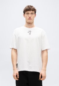 Λευκό βαμβακερό T-shirt με μικρό γκρι γραφικό στο στήθος, άνετη εφαρμογή, κοντά μανίκια και ετικέτα λογοτύπου στο πλάι του τελειώματος.