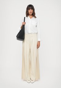 Beige plissert bukser med høy midje, kombinert med en hvit skjorte med knapper og svart totebag. Skoene er spisse og hvite.