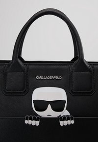 Musta käsilaukku, jossa on teksturoitu materiaali, kaksoiskahvat ja sarjakuvagrafiikka, joka kurkistaa reunan yli. Hopeinen logo korostaa grafiikan yläpuolella.