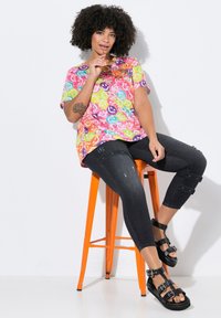 Haut à manches courtes multicolore avec des motifs vibrants, associé à un pantalon capri en denim foncé avec des détails déchirés. Sandales noires à lanières.