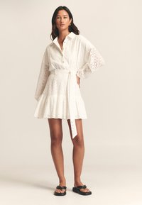 Bílé šaty ze síťoviny s límečkem, dlouhými rukávy a vrstvovou sukní. Mají pás s páskem, zapínání na knoflíky vpředu a charakteristický perforovaný vzor.