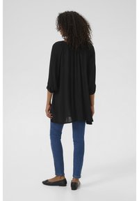 Blouse noire à manches longues avec une coupe ample et un dos froncé. Associée à un jean bleu et des ballerines noires, mettant en valeur une silhouette fluide.