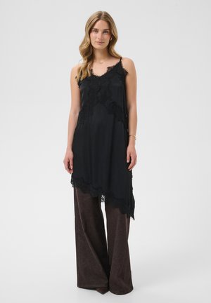 Femme debout, portant une tunique noire à bordure en dentelle sur un pantalon marron à jambes larges, avec les cheveux lâchés, un collier simple et un bracelet sur fond blanc.