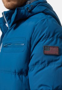 Blue puffer jacket with a zippered pocket on the chest and a textured American flag patch on the sleeve. Features a drawstring hood.
