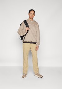 Beige hoodie met trekkoorden, gelaagd over een zwart shirt, gecombineerd met kaki broek en beige sneakers. Zwarte rugzak over één schouder geslagen.