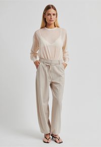 Lehký průsvitný broskvový top s dlouhým rukávem vrstvený přes béžové vysoké široké kalhoty s opaskem, doplněné černými plochými sandály.