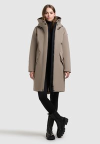 Long beige parka coat with a hood, featuring a zipper and snap button closure, front pockets, and a black accent zipper detail.