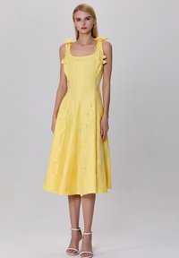 Robe jaune avec décolleté carré, bretelles spaghetti et accents en nœud ; design fluide mi-long en tissu doux avec une texture subtile.