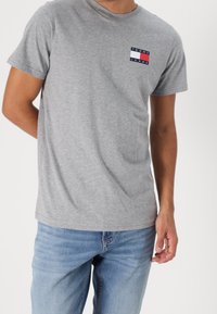 Hombre con una camiseta gris de manga corta Tommy Jeans y jeans azul claro, de pie contra un fondo blanco liso.