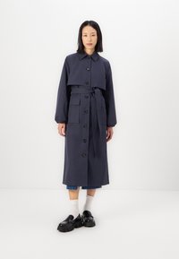 Trench coat di un blu scuro navy con uno strato superiore accorciato, chiusura con bottoni, cintura in vita e tasche. Presenta maniche lunghe e una finitura liscia.