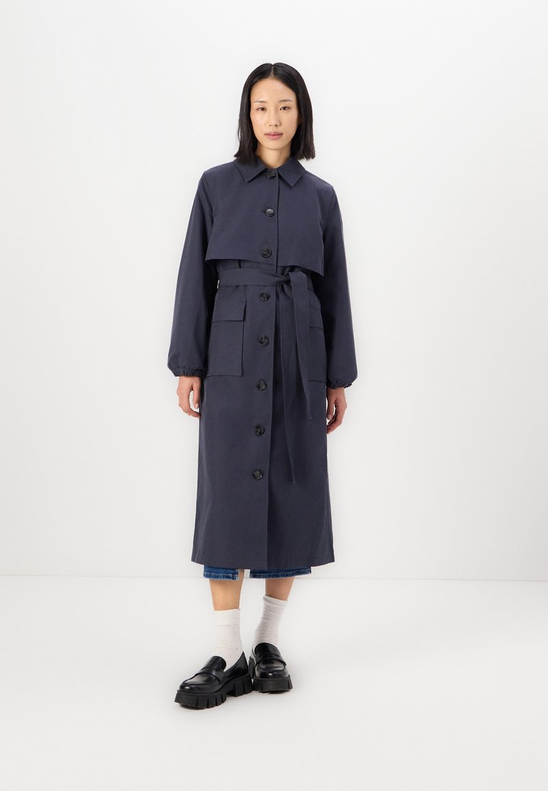 Trench coat di un blu scuro navy con uno strato superiore accorciato, chiusura con bottoni, cintura in vita e tasche. Presenta maniche lunghe e una finitura liscia.