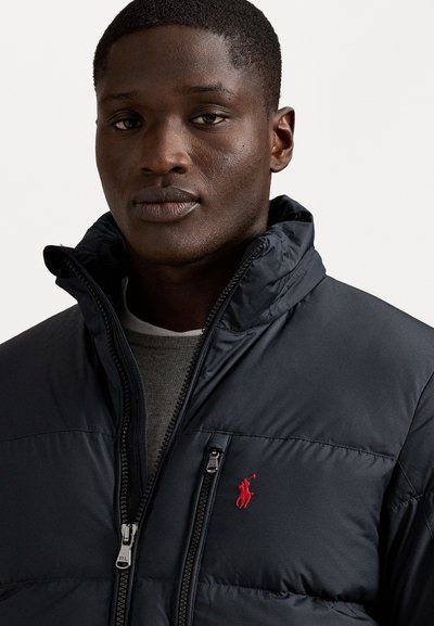 Black puffer jacket with a high collar, zip closure, and a red embroidered logo on the chest. Soft, quilted texture with zip pocket detail.