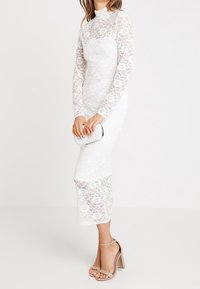 Weißes Spitzen-Midi-Kleid mit langen Ärmeln, floralen Mustern, hohem Kragen und durchsichtigen Akzenten. Kombiniert mit einer silbernen Clutch und beigen Riemchen-Heels.