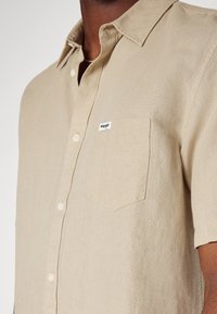 Camicia beige a maniche corte realizzata in tessuto testurizzato, con chiusura a bottoni frontale, colletto e taschino sul lato sinistro con etichetta del marchio.