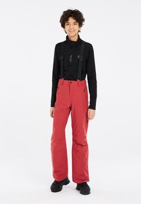 Pantaloni da sci rossi impermeabili con bretelle nere regolabili e tasche laterali, abbinati a un top nero a maniche lunghe e stivali neri.