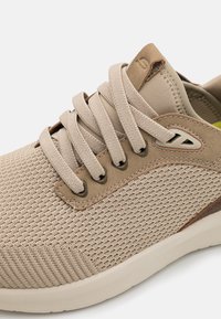 Zapatilla deportiva beige con parte superior de malla texturizada, cordones planos y un acento de cuero suave. Punta redonda y suela acolchada para mayor comodidad.