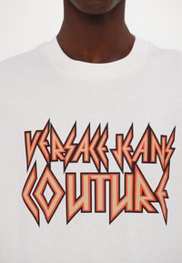 Bela bomba majica iz bombaža z drznim, ostrim napisom v oranžni in črni barvi, ki pravi "VERSACE JEANS COUTURE." Klasičen kroj s krožnim izrezom.