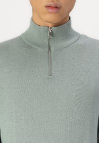 Light green zip-up sweater with a high collar. Made of a soft knit material featuring a smooth texture and subtle sheen.
