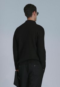 Schwarzer gerippter Pullover mit Kragen, in entspannter Passform. Das Model hält eine schwarze Leder-Clutch und trägt eine Sonnenbrille.