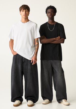 To modeller bærer oversized t-shirts: en hvid og en sort. Begge har dem på med mørke, vide jeans og lyse sneakers. Simpelt design, afslappet pasform.