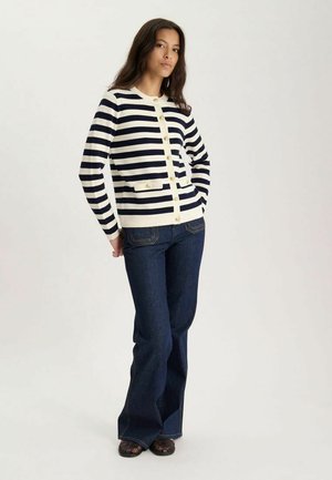 Femme debout portant un cardigan boutonné rayé bleu marine et blanc, un jean évasé bleu foncé et des chaussures tissées foncées sur un fond clair.