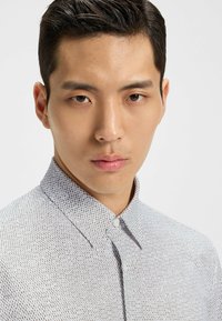 Biała koszula z guzikami, z geometrycznym ciemnym wzorem, z klasycznym kołnierzykiem i dopasowanym krojem. Gładki materiał, minimalistyczny design.