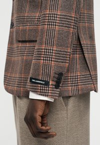 Manche de blazer à carreaux marron et noir avec boutons noirs et étiquette Karl Lagerfeld au poignet, portée sur un pantalon beige et un poignet de chemise blanche.