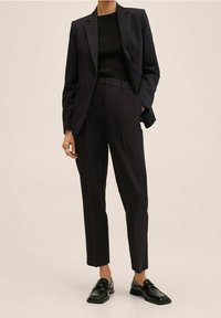 Costume bleu marine avec blazer cintré et pantalon sur mesure, associé à un haut noir côtelé et des mocassins noirs brillants. Tissu lisse, silhouette nette.