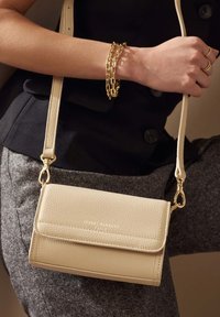 Beige leather crossbody bag with a textured finish, gold-tone hardware, and a magnetic flap closure, worn on a black outfit.