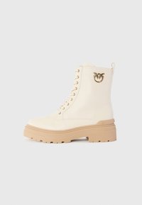 Crème-kleurige leren veterschoen met een dikke beige zool en een gouden vogel-embleem aan de zijkant, gefotografeerd op een witte achtergrond.