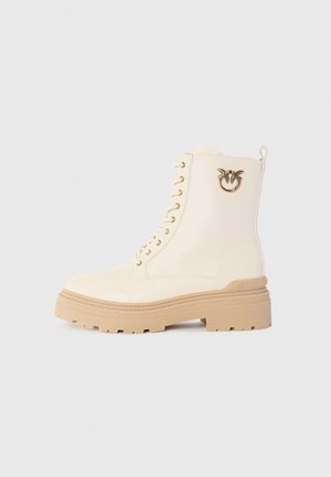 Crème-kleurige leren veterschoen met een dikke beige zool en een gouden vogel-embleem aan de zijkant, gefotografeerd op een witte achtergrond.