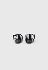 Ballerines en cuir noir avec un bout arrondi, une bride de cheville et un détail de nœud décoratif à l'arrière. Texture lisse avec un éclat subtil.