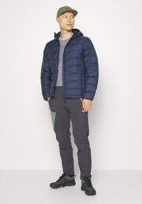Giubbotto imbottito blu navale con cappuccio, maglietta grigia, pantaloni grigi con accenti verdi e arancioni, e scarpe da trekking nere. Il modello è in piedi contro uno sfondo semplice.