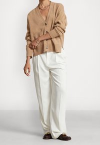 Beige stickad cardigan med v-ringning, knäppning framtill och ribbade muddar, kombinerad med vita veckade byxor och mörka loafers.