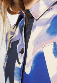 Lähikuva sinisestä, purppurasta ja valkoisesta kuviollisesta takista, jossa on mustia nappeja, kaulus ja tasku, jonka päällä on musta vaate ja osittain näkyvissä pitkät ruskeat hiukset.