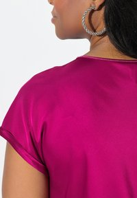Top en satin magenta à manches courtes, texture lisse et éclat subtil ; présente un col rond avec un détail de couture décoratif.