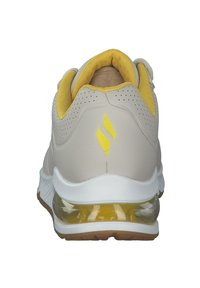 Vaaleanbeige urheilukengät, joissa on keltainen pehmustettu kaulus, takana rei'itetty muotoilu, kirkas ilmasolu ja keltainen korostuslogo.