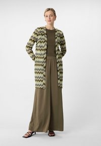 Długa oliwkowozielona sukienka z teksturowanej tkaniny, warstwowa pod wzorzystym kardiganem z zygzakowymi pasami w odcieniach zieleni, czerni i bieli. Czarne sandały na paski.