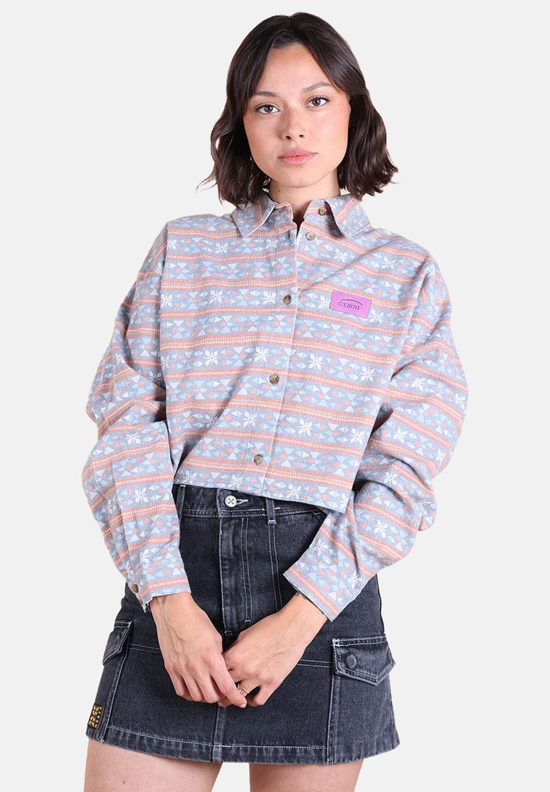 Femme aux cheveux foncés mi-longs, portant une chemise à motifs boutonnée et une jupe en denim noire, se tenant les mains jointes devant elle.