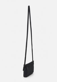 Melna crossbody soma, kas izgatavota no izturīga auduma, ar garu regulējamu siksnu, taisnstūra formā un ar rāvējslēdzēju aizveramu kabatu priekšpusē.