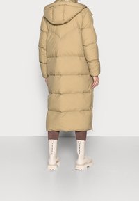 Abrigo largo beige tipo plumón con capucha, con secciones acolchadas horizontales. Combinado con botas crema hasta la rodilla. Textura suave.