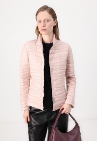 Μαλακή ροζ παλτό με ψηλό γιακά, κατακόρυφη ραφή και κλείσιμο με κουμπιά. Φοριέται πάνω από ένα μαύρο μπλουζάκι και συνδυάζεται με σκούρα δερμάτινα παντελόνια.