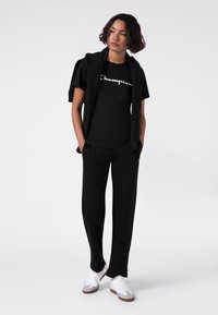 Schwarz Baumwoll-T-Shirt mit weißem "Champion"-Logo, kombiniert mit schwarzen Jogginghosen. Das Model trägt eine schwarze Jacke lässig über den Schultern und weiße Schuhe.