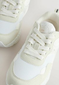 White sneakers with textured fabric and suede accents. Lightweight design features rounded toe, flat laces, and logo on the tongue.