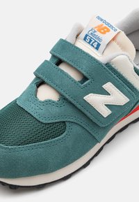 Svetle zelené semišové a síťované tenisky s dvojitými suchými zipy, bílými akcenty a červenými detaily. Na jazyku je označení New Balance. Klasický design.
